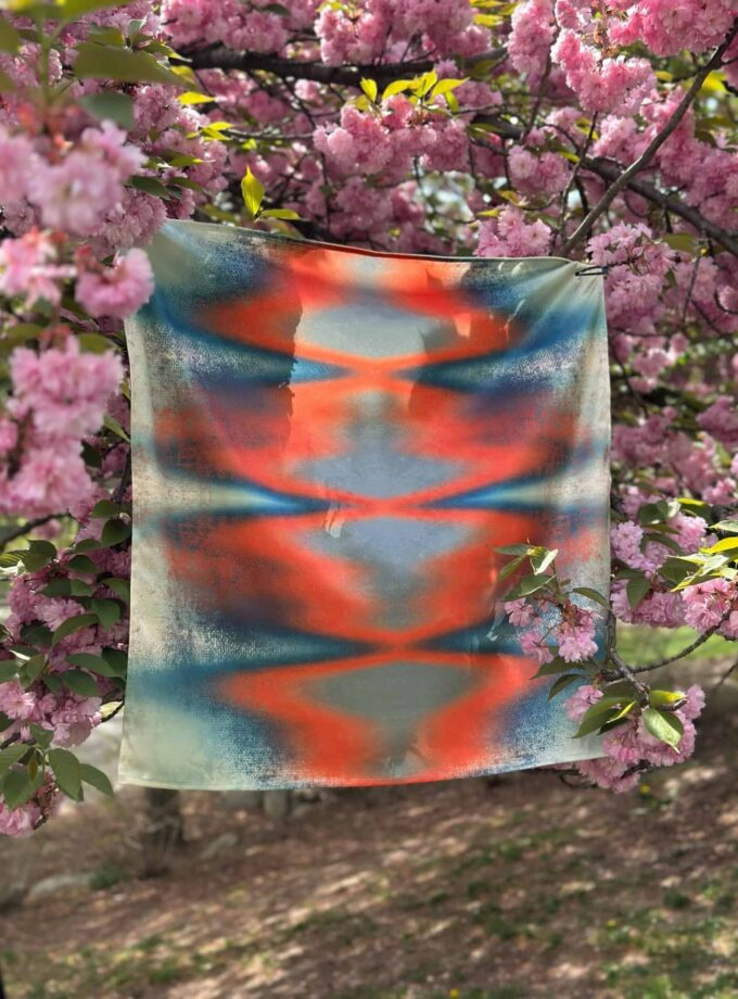 Vibrant abstract art scarf hanging on a branch of blooming pink cherry blossoms, showcasing colorful, handcrafted textile designs inspired by nature and artistic expression.