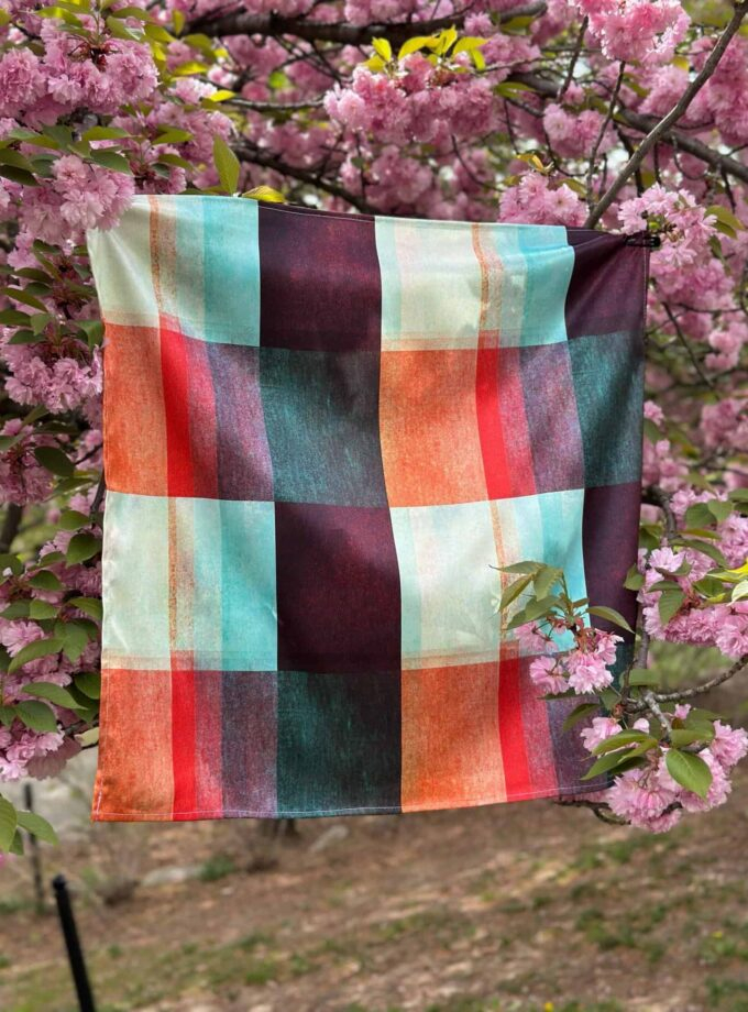 Vibrant checkered cloth hanging on pink cherry blossom tree, showcasing colorful table linens for outdoor gatherings, picnic, or dessert presentation, perfect for springtime and garden settings.