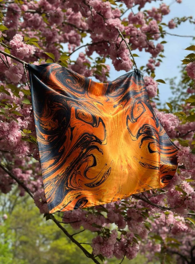 Vibrant orange and black marbled silk scarf hanging on cherry blossom tree branch, showcasing unique textile design amidst blooming pink flowers.