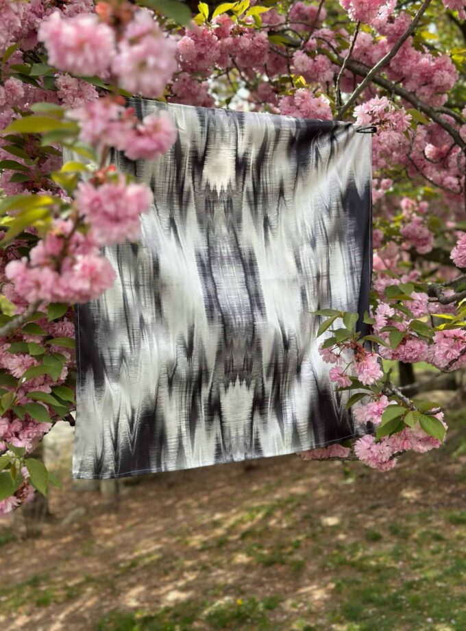 Vibrant pink cherry blossoms surrounding a black and white abstract textile art piece hanging on a tree branch in a garden.
