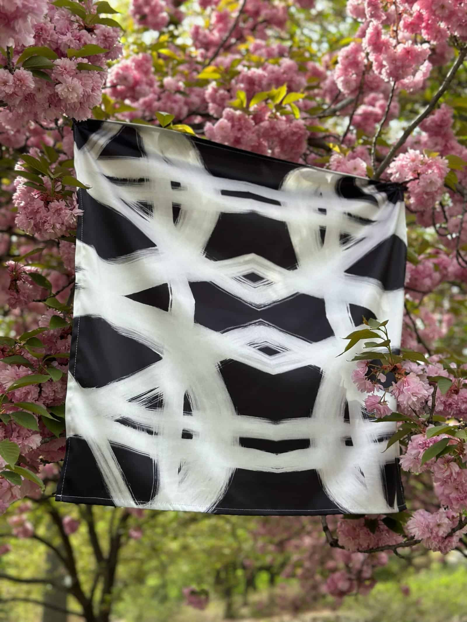 Vibrant black and white abstract patterned scarf hanging among pink cherry blossoms. Elegant accessory with artistic design, perfect for spring fashion and outdoor style.