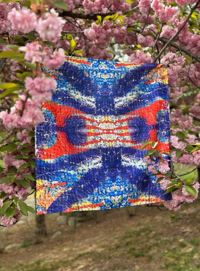 Vibrant abstract textile art hanging on blooming pink cherry blossom tree in spring.