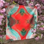 Vibrant abstract patterned scarf hanging on a branch with pink cherry blossoms in the background, featuring bold colors and artistic design for fashionable accessories.