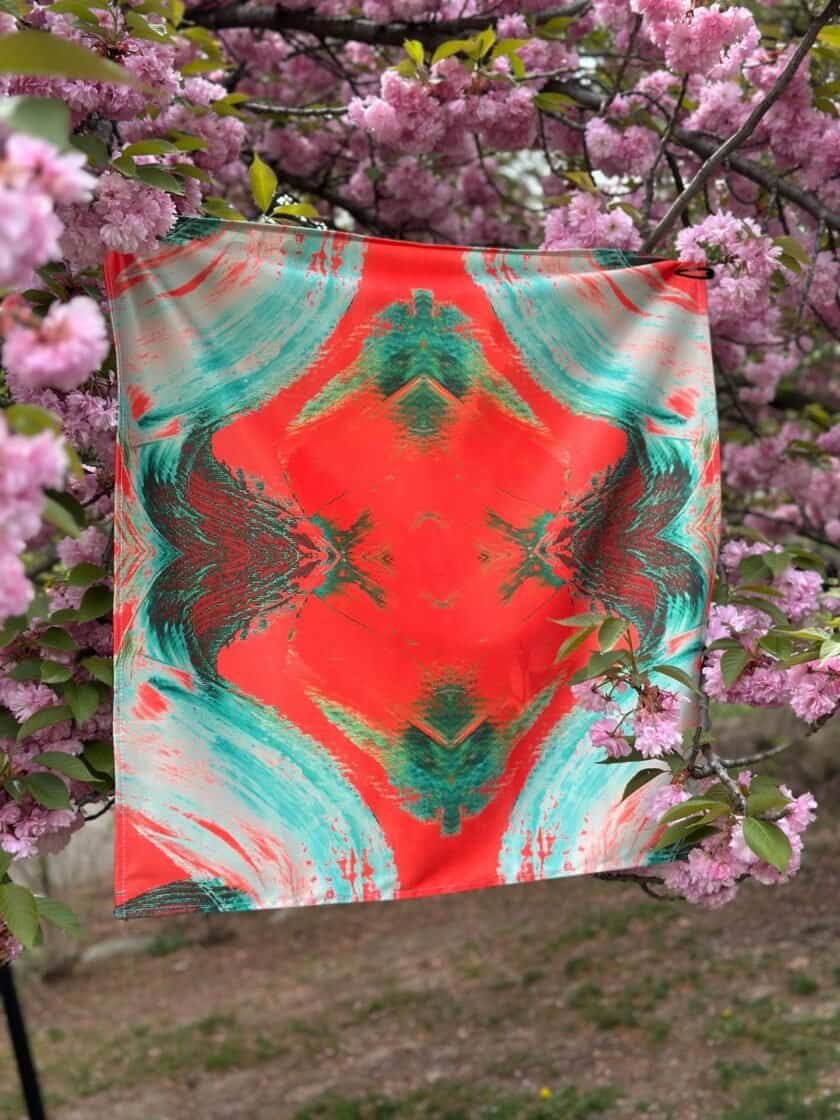 Vibrant abstract patterned scarf hanging on a branch with pink cherry blossoms in the background, featuring bold colors and artistic design for fashionable accessories.