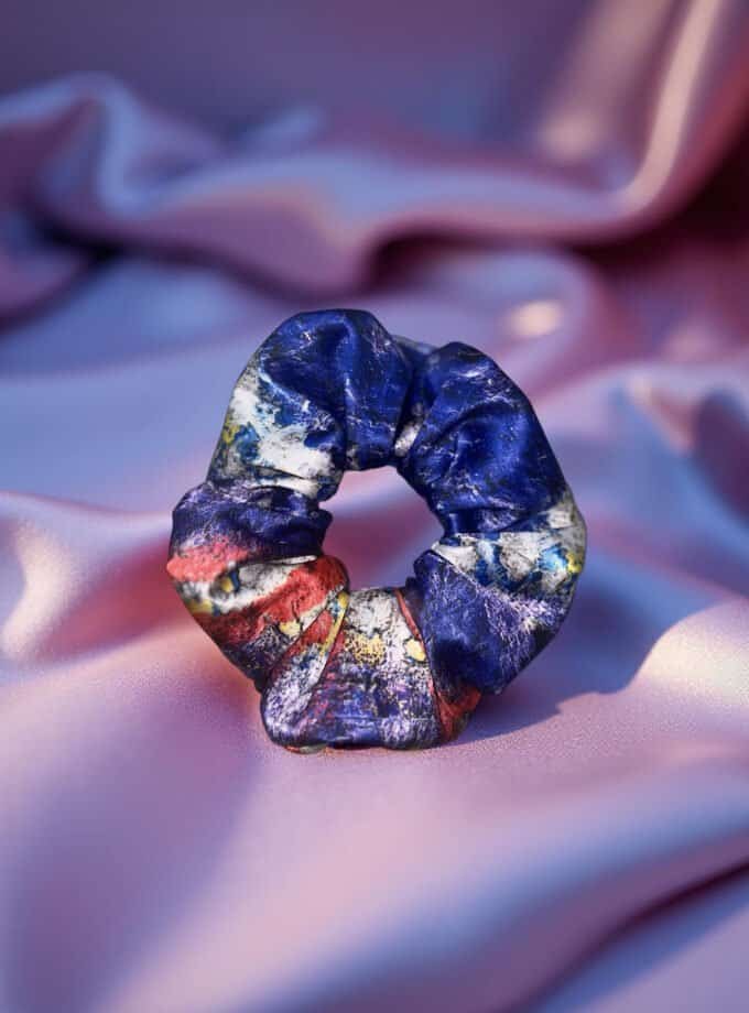 Vibrant abstract scrunchie with a mix of blue, red, white, and yellow colors, set against soft pink satin fabric, showcasing artistic and colorful hair accessories from Confiture.