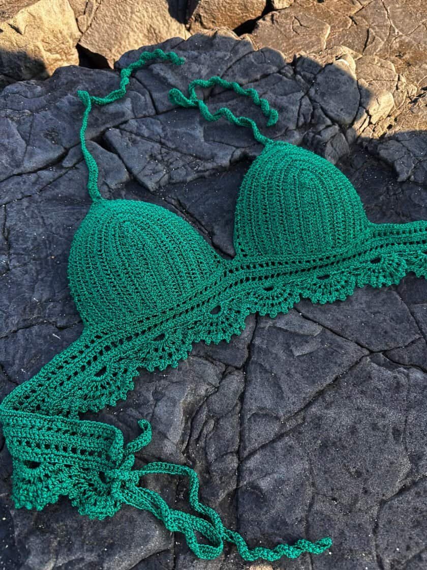 Bright green crocheted bikini top with scalloped edges, laid on dark, textured rocks. Perfect for summer beachwear, showcasing handmade swimwear craftsmanship and vibrant color. Ideal for seaside lifestyle imagery.