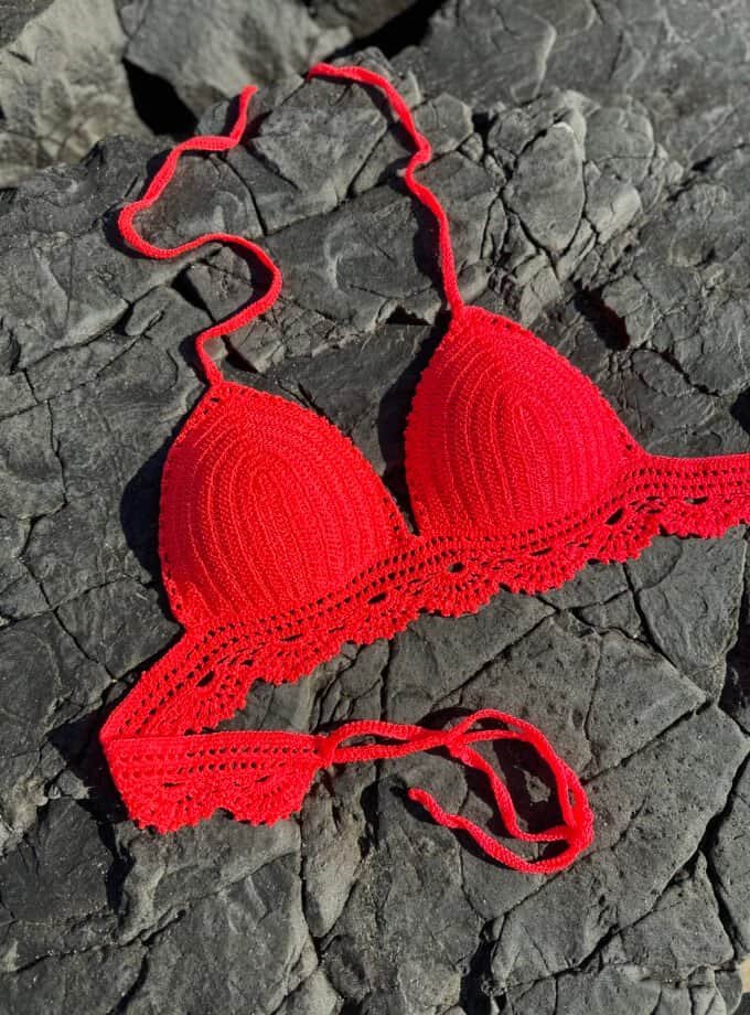 Vibrant red crochet bikini top placed on dark, rugged rocks, highlighting handmade swimwear designed for beachwear fashion and summer style.