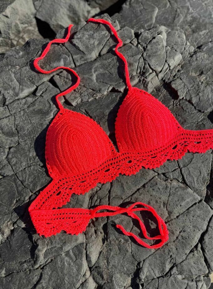 Vibrant red crochet bikini top placed on dark, rugged rocks, highlighting handmade swimwear designed for beachwear fashion and summer style.