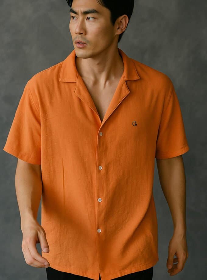 Orange shirt with a small embroidered logo on the chest, modern men's fashion, stylish casual wear, lightweight linen fabric, short-sleeve button-up, summer clothing, fashion photography, grey textured background.