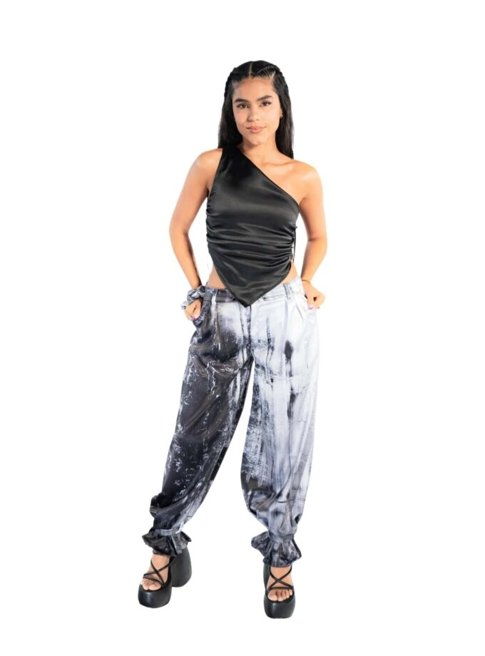 Stylish young woman wearing a black satin one-shoulder top and black-and-white tie-dye paint splatter baggy pants with platform heels for a trendy fashion look.