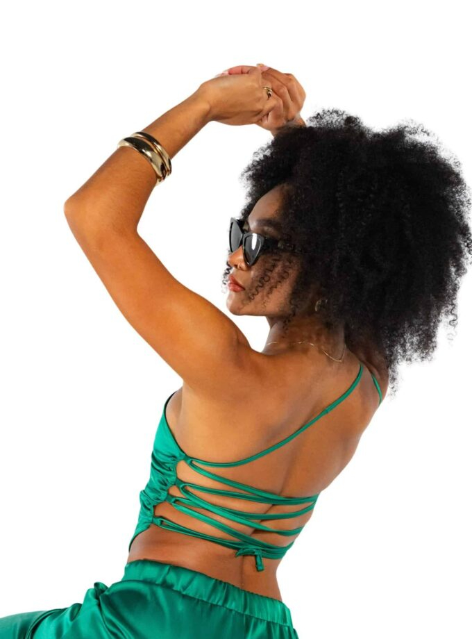 Vibrant woman with natural afro hair in stylish green satin outfit, wearing sunglasses, jewelry, and posing confidently against a white background, showcasing summer fashion and lifestyle.