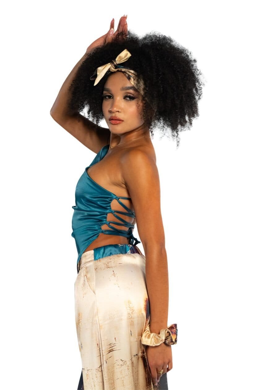 Bright young woman with Afro hair wearing a satin blue top and cream-colored tie-dye pants, accessorized with a beige silk scarf headband, stylish jewelry, and a scrunchie, posing confidently.