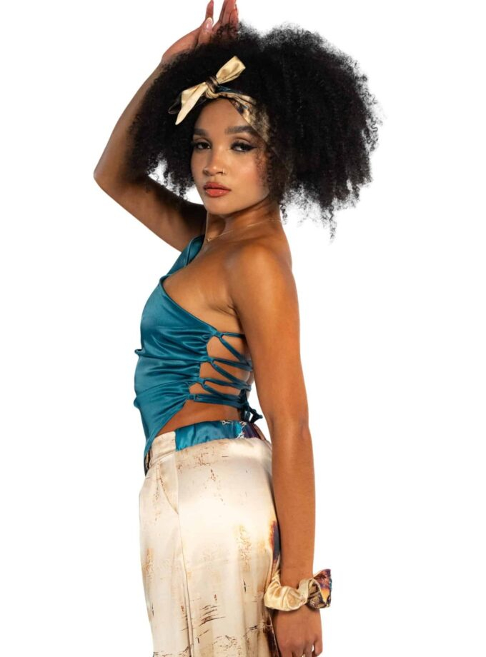 Bright young woman with Afro hair wearing a satin blue top and cream-colored tie-dye pants, accessorized with a beige silk scarf headband, stylish jewelry, and a scrunchie, posing confidently.