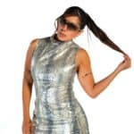 Elegant woman in metallic snake print sleeveless dress with black tights, wearing stylish sunglasses, posing confidently with her long hair, on a plain white background.