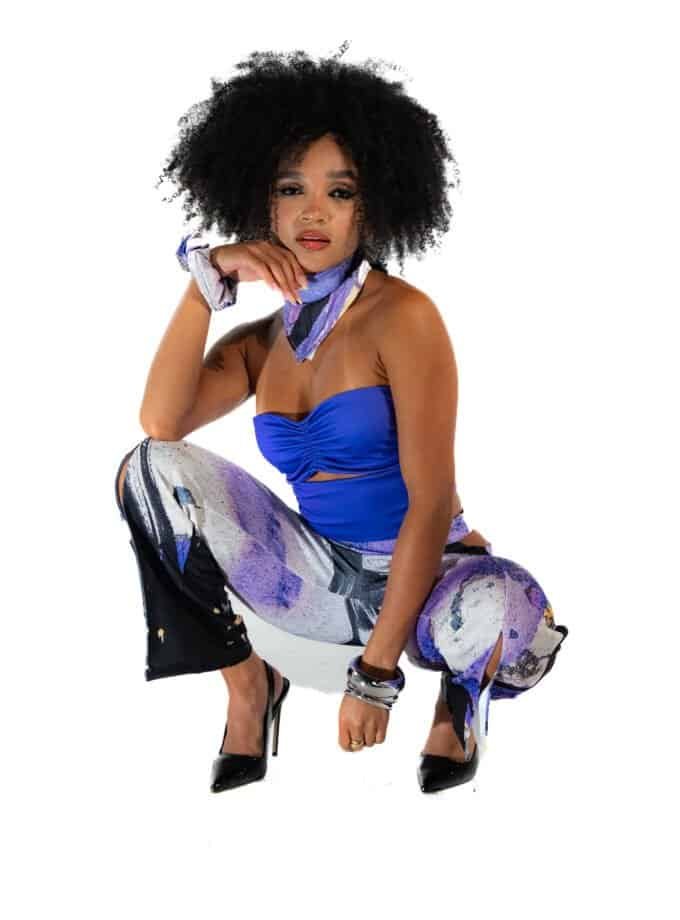 Vibrant stylish woman in a blue strapless top and colorful abstract print pants, squatting with high heels, showcasing fashion and confidence, against a clean white background.