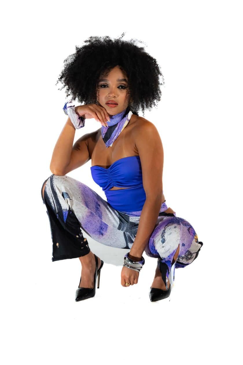 Vibrant stylish woman in a blue strapless top and colorful abstract print pants, squatting with high heels, showcasing fashion and confidence, against a clean white background.