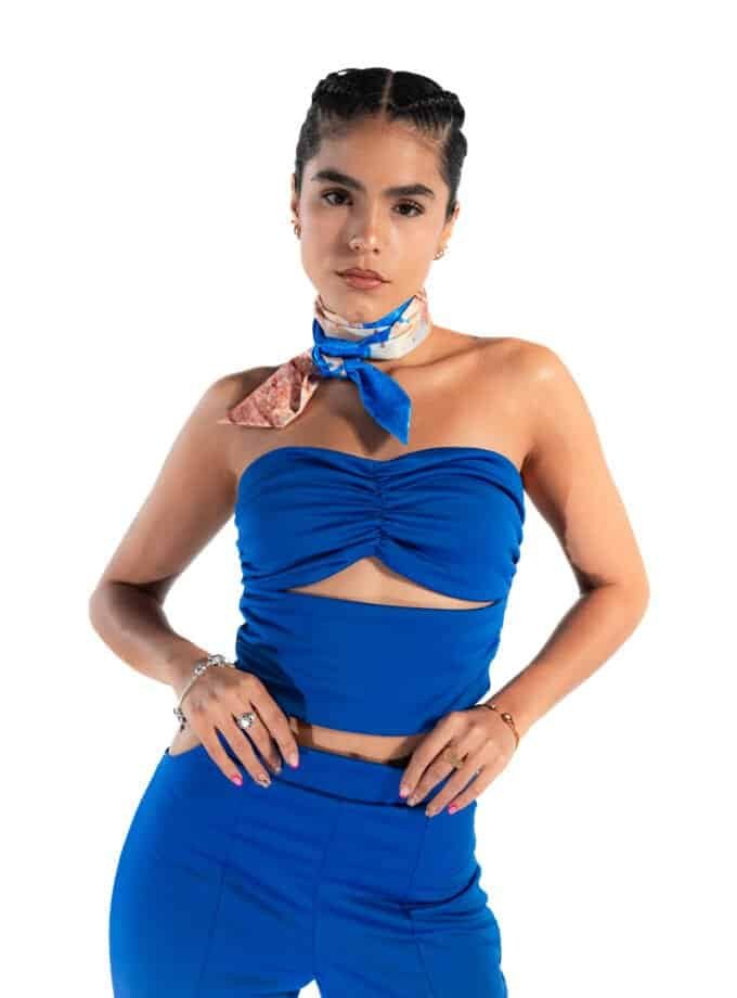 Bright woman in a stylish blue crop top and matching high-waisted pants, accessorized with jewelry and a colorful scarf tied around her neck, showcasing modern fashion and confidence.