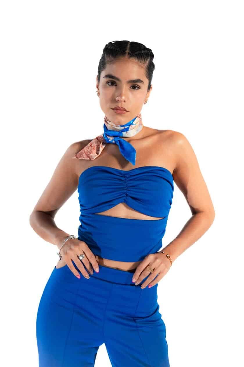 Bright woman in a stylish blue crop top and matching high-waisted pants, accessorized with jewelry and a colorful scarf tied around her neck, showcasing modern fashion and confidence.