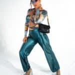 Shiny teal satin pants and matching crop top with digital print, stylish futuristic sunglasses, and platform heels, model poses confidently in a fashion-forward look for Confiture NYC.