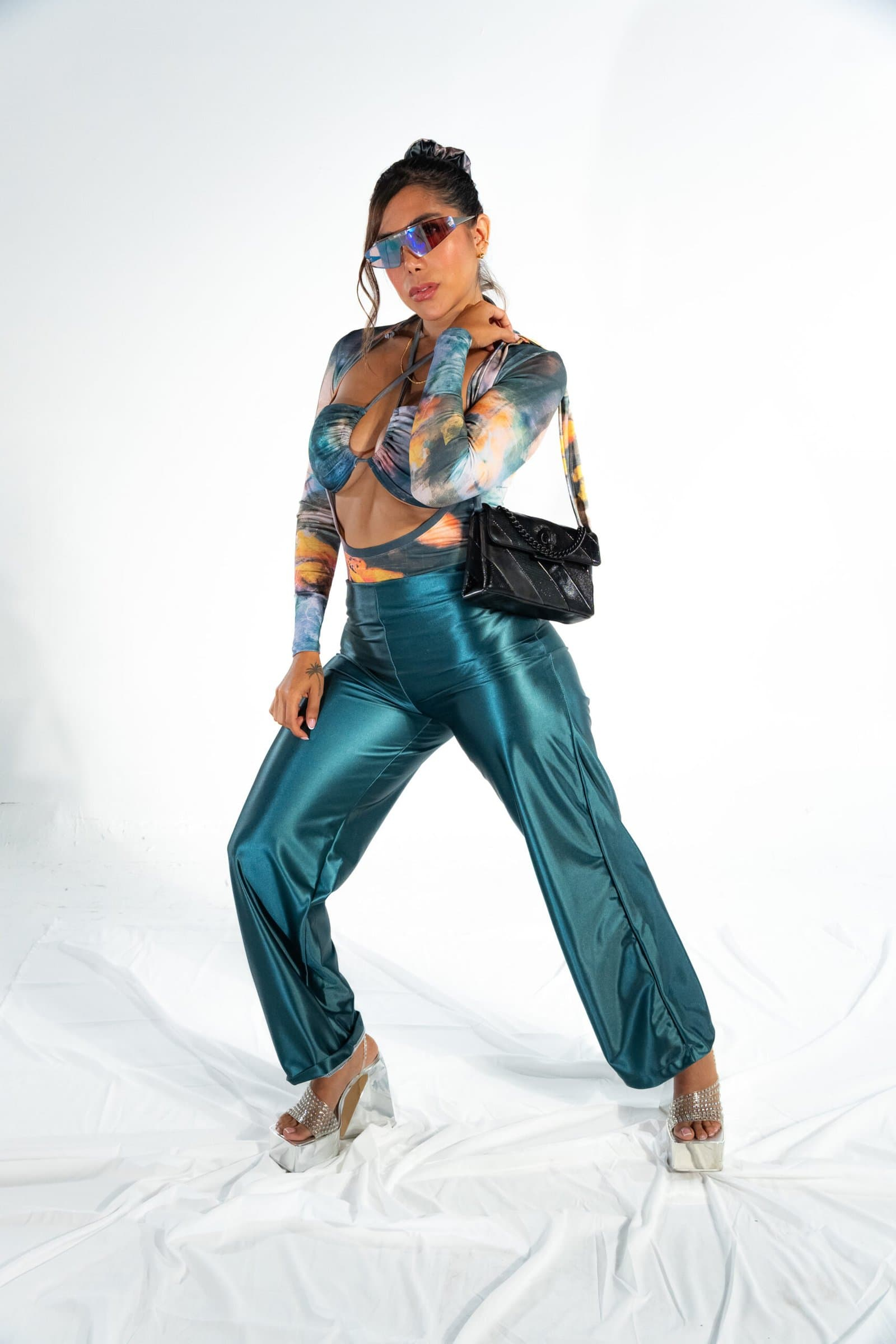 Shiny teal satin pants and matching crop top with digital print, stylish futuristic sunglasses, and platform heels, model poses confidently in a fashion-forward look for Confiture NYC.