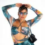 Vibrant woman in futuristic sunglasses wearing a colorful, cutout top and shiny pants, capturing a bold, fashion-forward look with a stylish handbag for a modern, trendy aesthetic.
