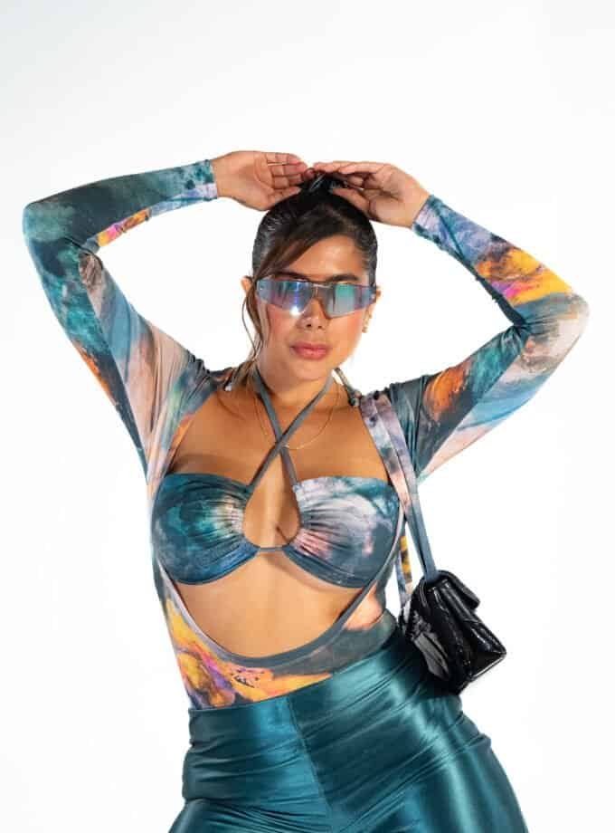 Vibrant woman in futuristic sunglasses wearing a colorful, cutout top and shiny pants, capturing a bold, fashion-forward look with a stylish handbag for a modern, trendy aesthetic.