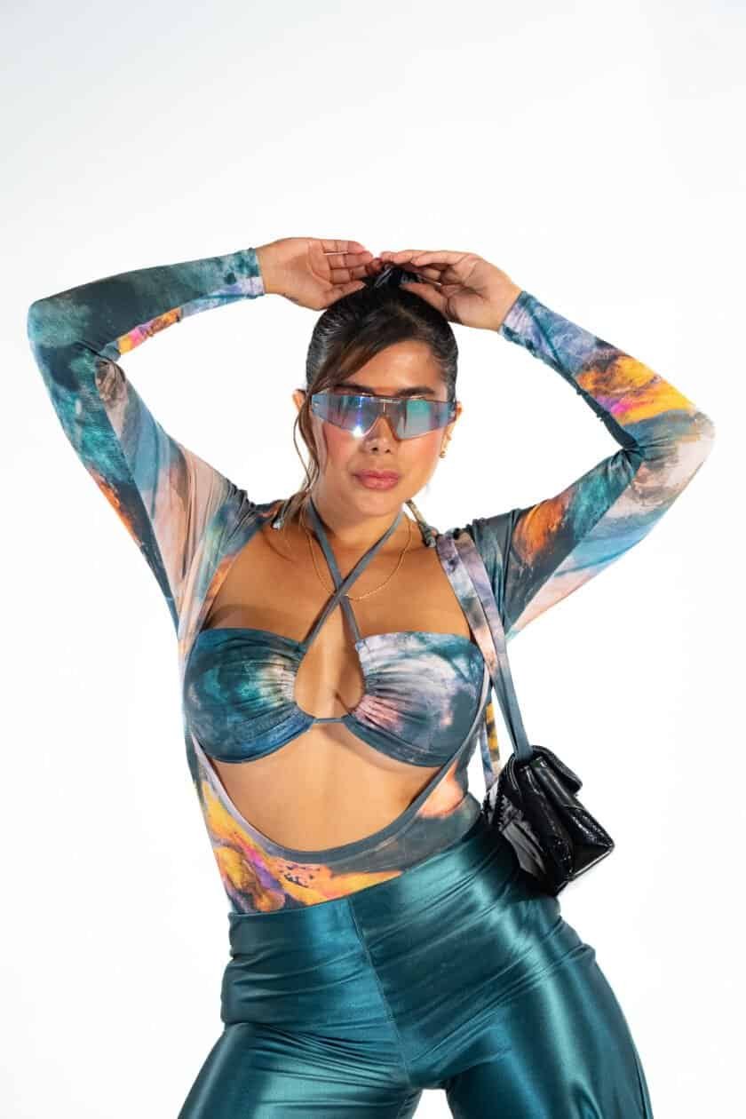 Vibrant woman in futuristic sunglasses wearing a colorful, cutout top and shiny pants, capturing a bold, fashion-forward look with a stylish handbag for a modern, trendy aesthetic.