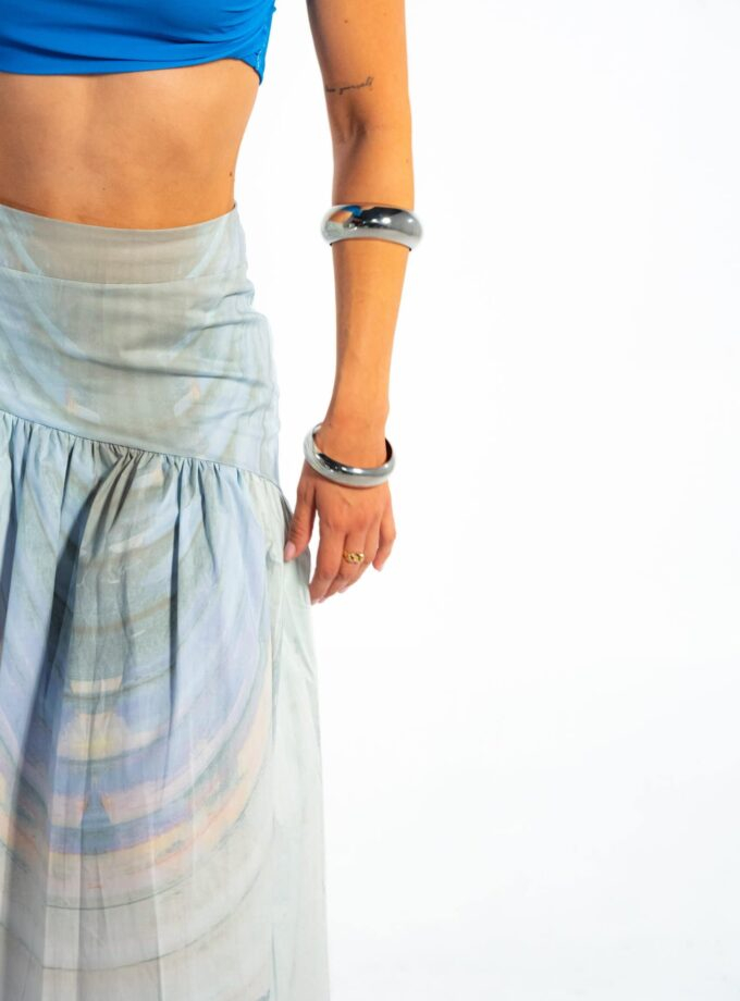 Elegant woman wearing metallic bangles, stylish pastel skirt, and vibrant blue top, showcasing contemporary fashion accessories against a minimal white background.