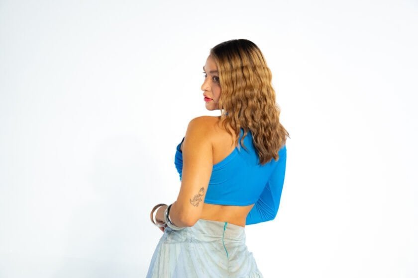 Blue one-shoulder crop top worn by woman with wavy hair, side profile, studio shot, featuring tattoo and jewelry, fashion and beauty, vibrant and stylish image.