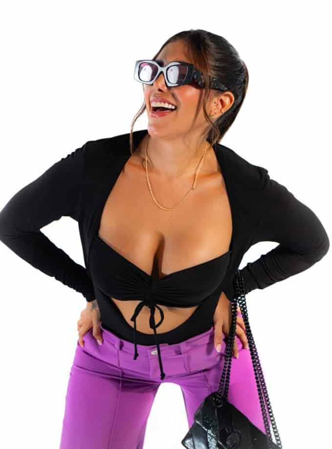 "Woman laughing wearing 3D glasses, black crop top, and purple pants, showcasing playful fashion and vibrant style, with a black purse and jewelry, on a white background.".