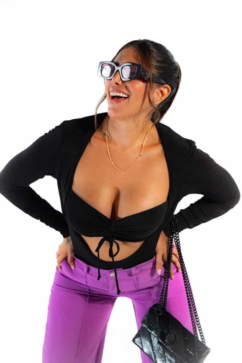 "Woman laughing wearing 3D glasses, black crop top, and purple pants, showcasing playful fashion and vibrant style, with a black purse and jewelry, on a white background.".