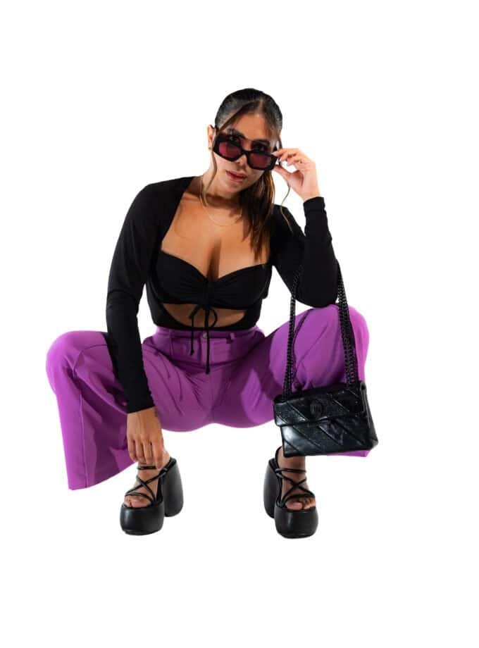 Vegan fashion model wearing trendy purple pants, black crop top, platform heels, and stylish sunglasses, holding a black handbag, showcasing street style and contemporary fashion trends.