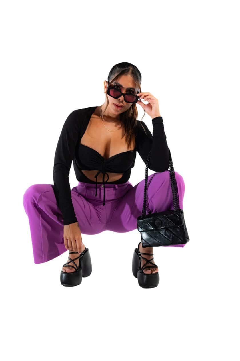 Vegan fashion model wearing trendy purple pants, black crop top, platform heels, and stylish sunglasses, holding a black handbag, showcasing street style and contemporary fashion trends.