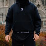 Black hoodie with white logo and Latin inscription, casual streetwear fashion, outdoor urban background.