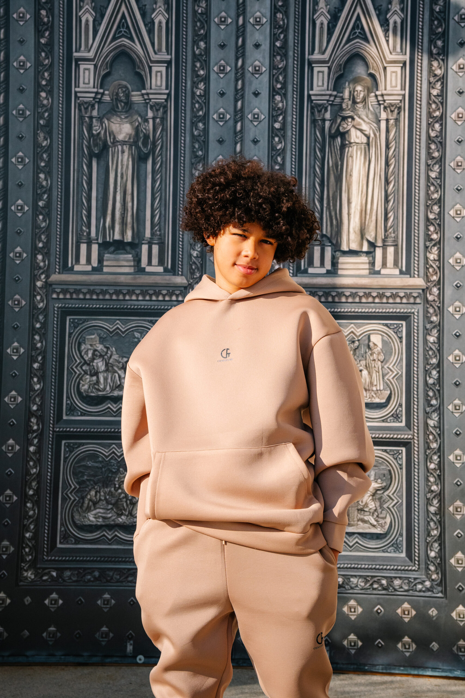 Comfortable beige hoodie and joggers set from Confiture NYC, featuring minimal branding, perfect for casual or urban streetwear looks, photographed outdoors against an ornate architectural background.