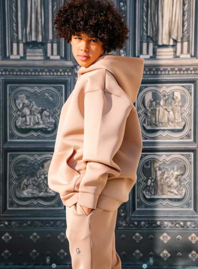 A young woman with curly hair in a beige hoodie and matching joggers standing against an ornate, historical architectural background. Stylish athleisure outfit in soft neutral tones.