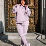 1. Woman in pastel lilac hoodie and joggers standing in front of ornate gold and black religious statue doorway in urban setting.