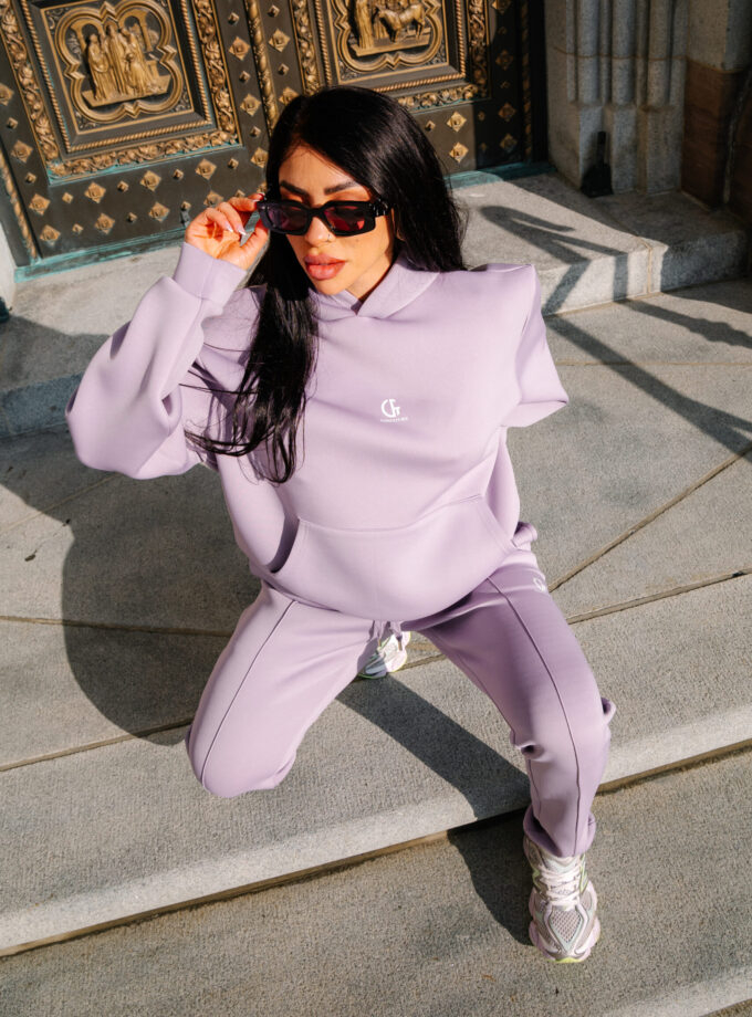 Vibrant woman in lilac athleisure squatting outdoors in front of ornate golden doors, wearing black sunglasses, showcasing stylish casual fashion for modern streetwear trends.