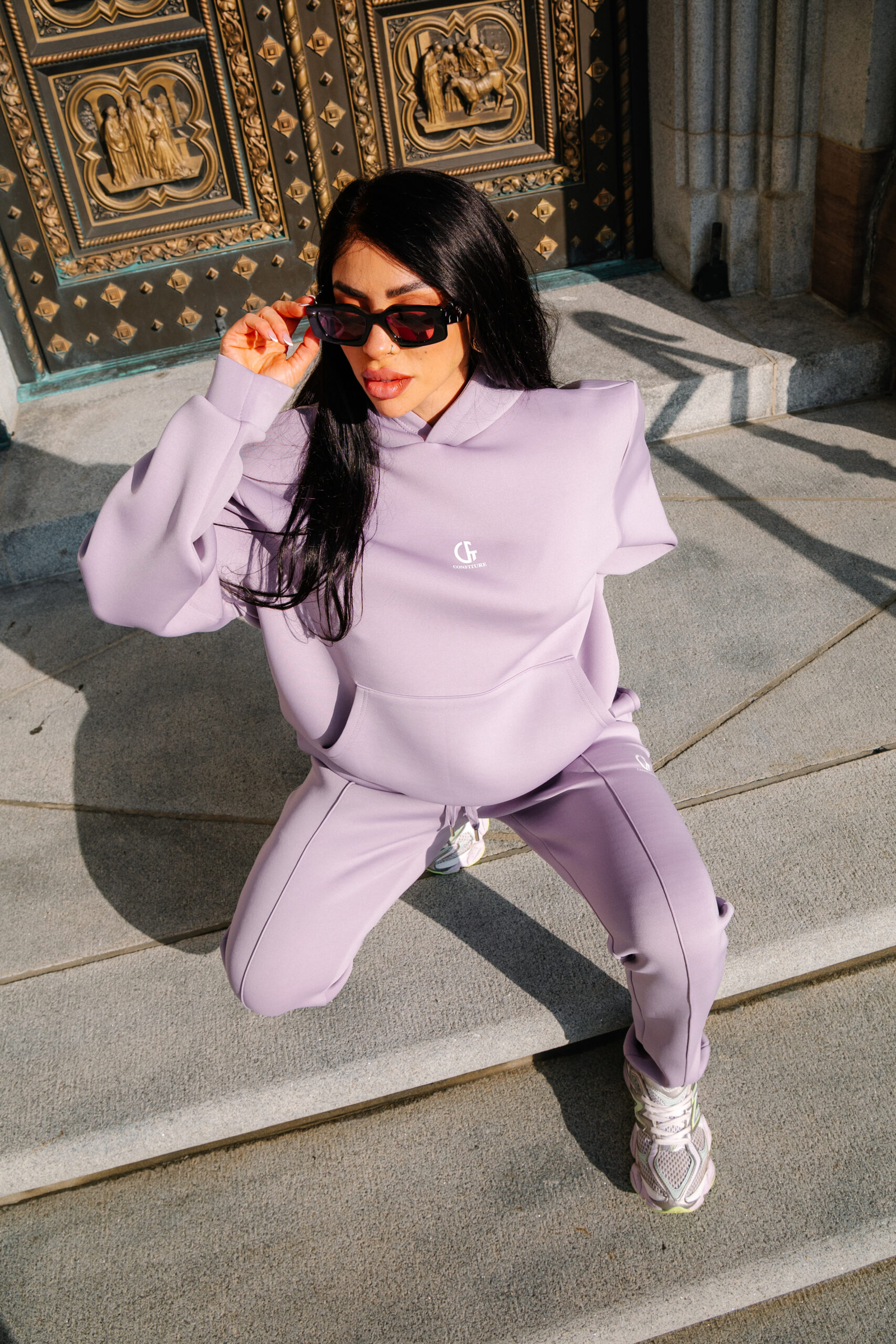 Vibrant woman in lilac athleisure squatting outdoors in front of ornate golden doors, wearing black sunglasses, showcasing stylish casual fashion for modern streetwear trends.