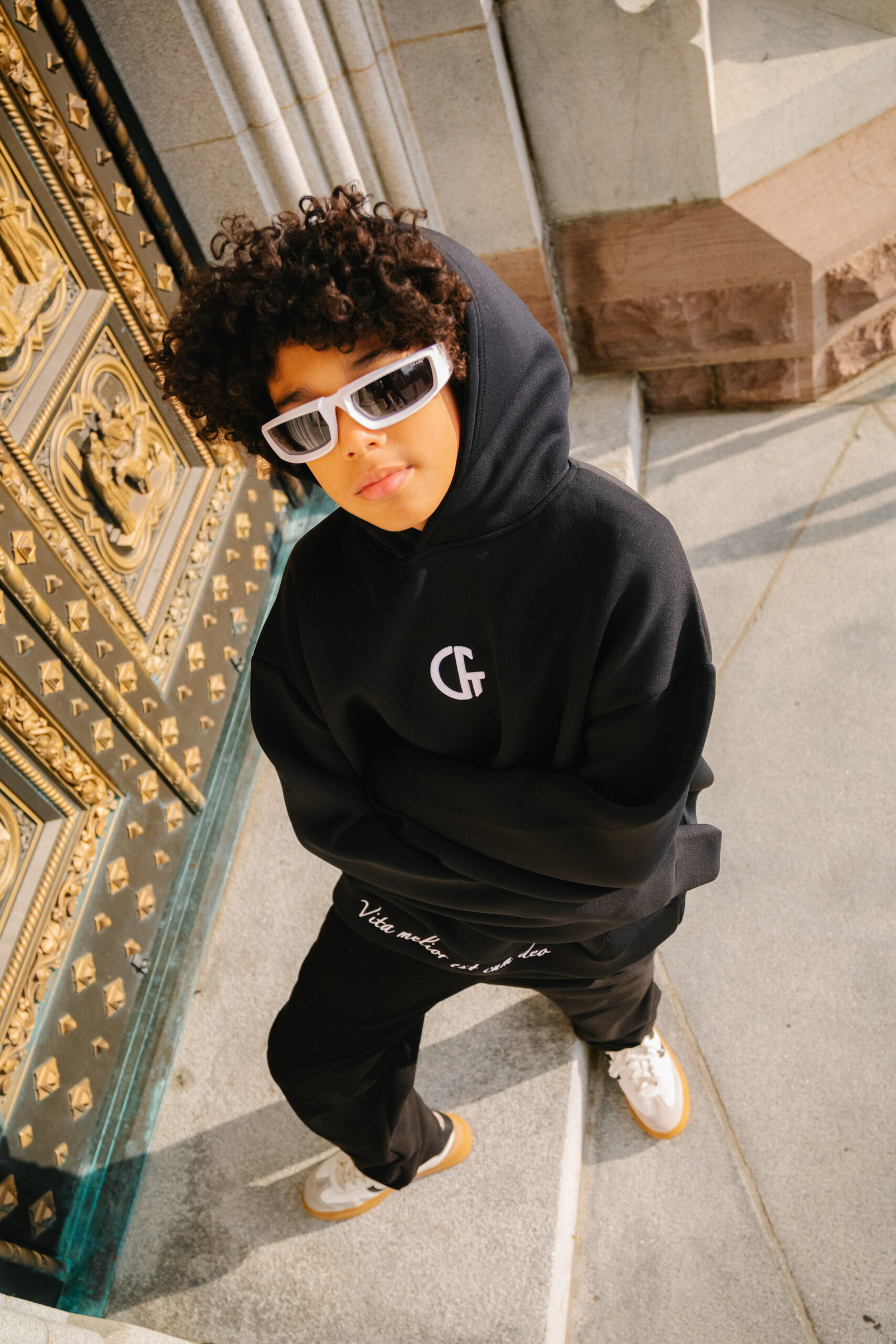 1. Boy wearing black hoodie and stylish sunglasses standing near ornate golden gate in an urban setting.