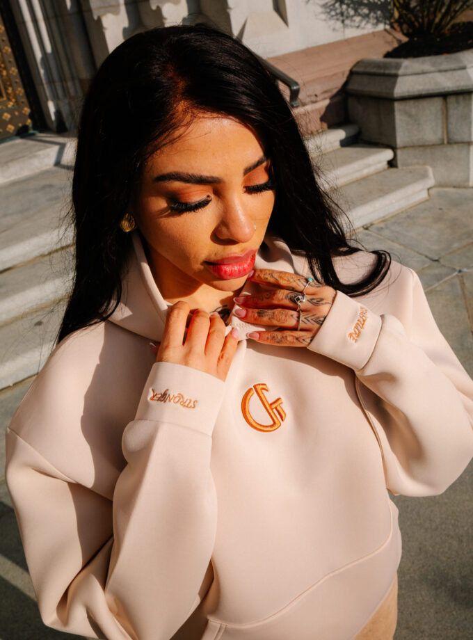 1. Close-up of a woman wearing a beige hoodie with Confiture logo, showcasing vibrant makeup and tattoos, standing outdoors with sunlight highlighting her face and outfit.