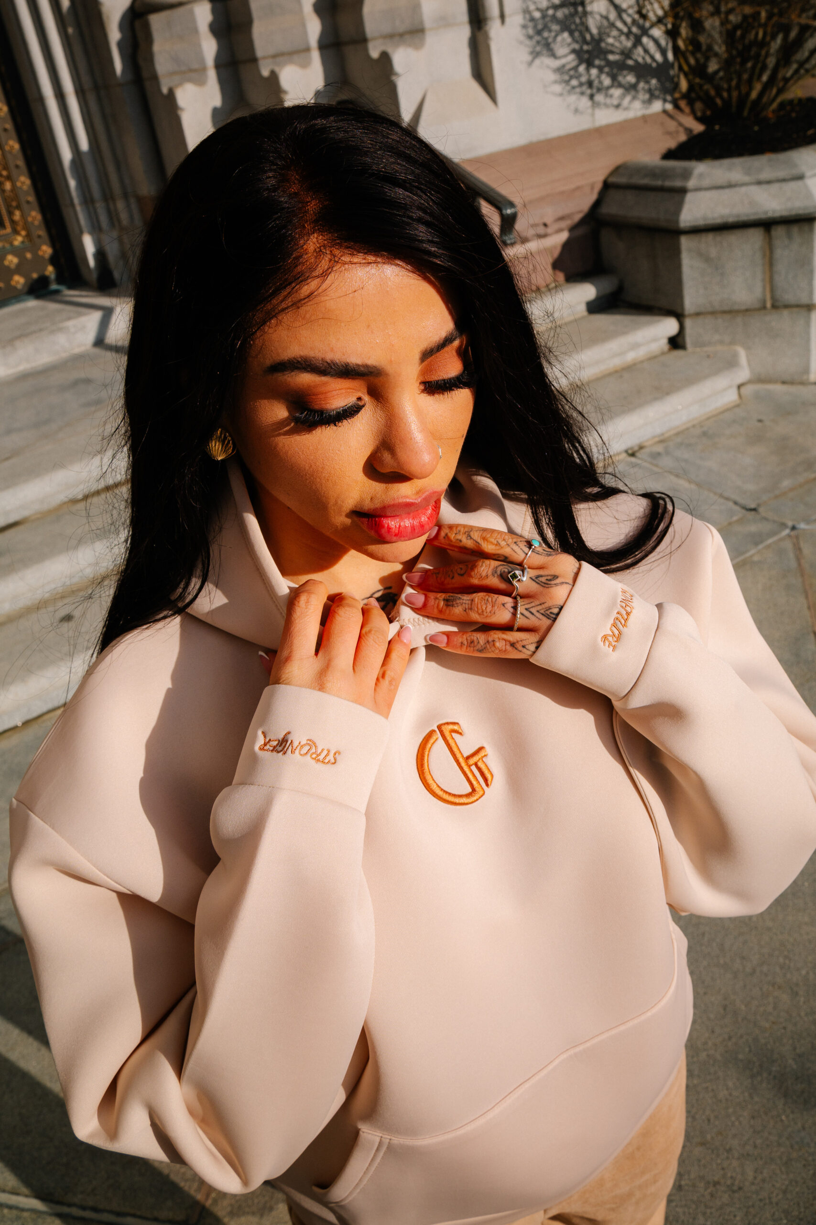 1. Close-up of a woman wearing a beige hoodie with Confiture logo, showcasing vibrant makeup and tattoos, standing outdoors with sunlight highlighting her face and outfit.