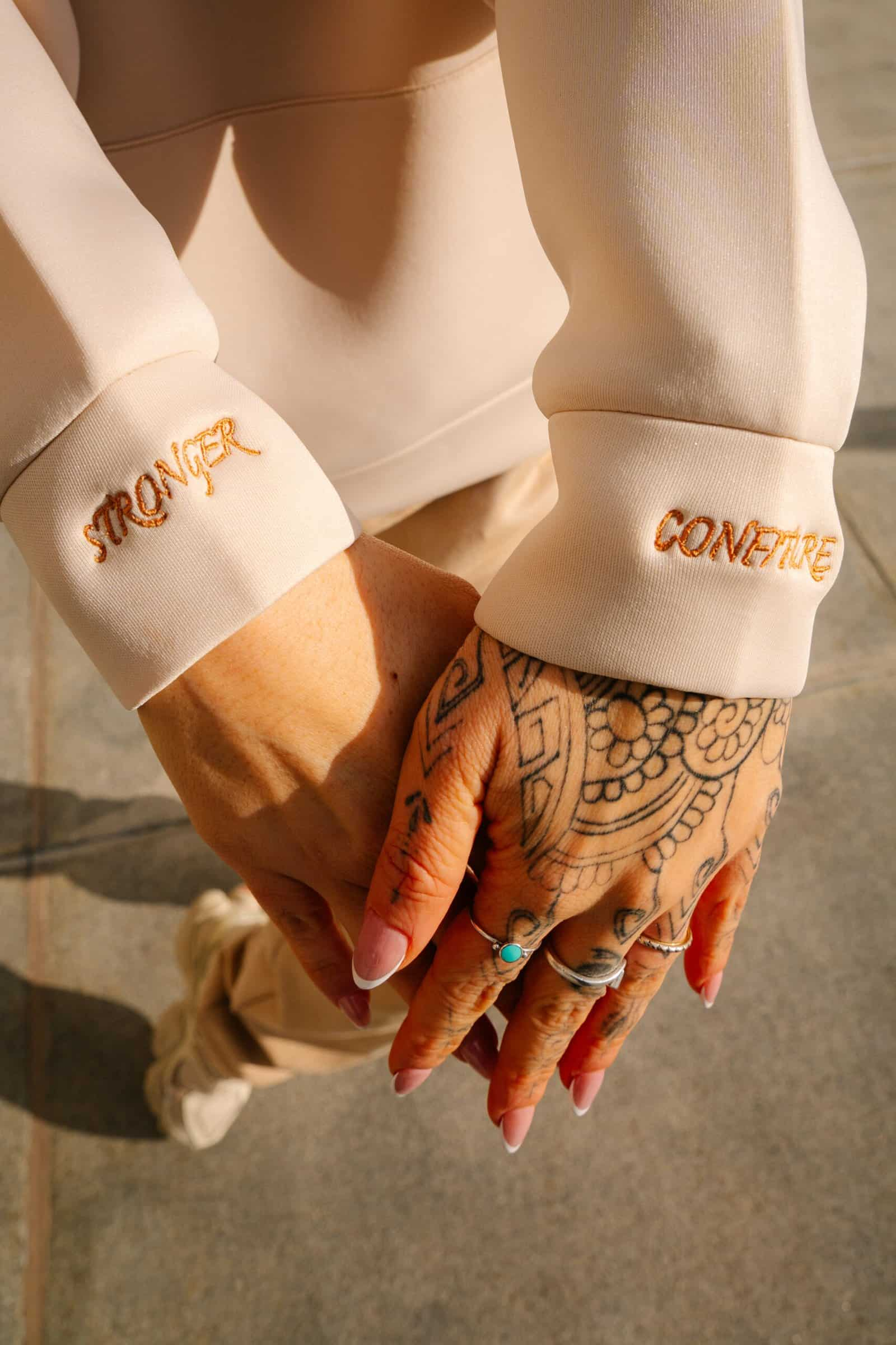 Elegant hands with tattoos and rings, wearing cream-colored sleeves embroidered with "Stronger" and "Confiture," symbolize strength and artisanal craftsmanship, showcasing a stylish and empowering vibe.