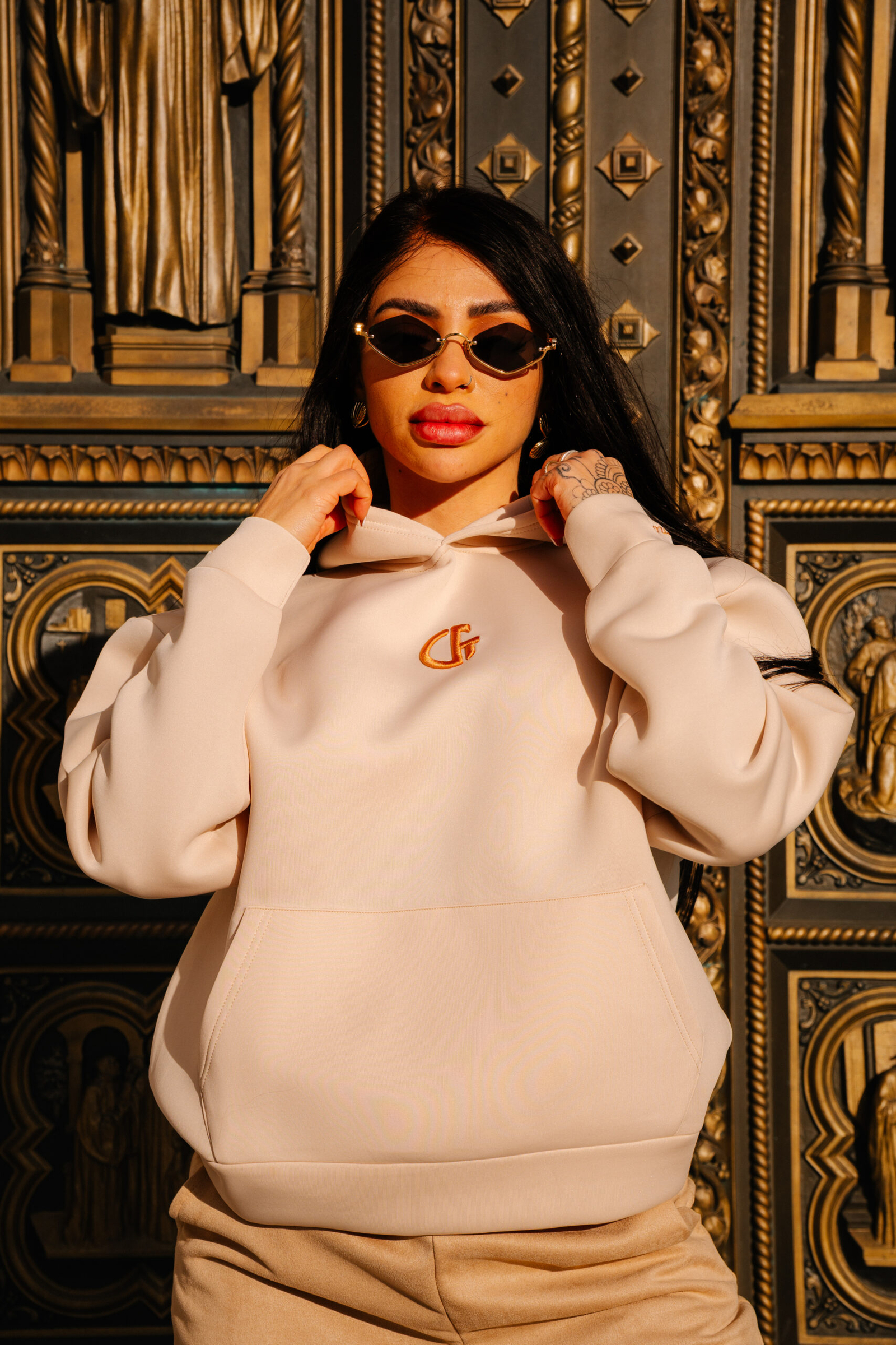 Bright young woman wearing a beige hoodie and stylish sunglasses at Confiture NYC, showcasing luxury fashion and premium streetwear in an ornate, gold-adorned setting.