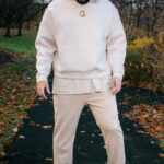 A man wearing a cream-colored hoodie and sweatpants, standing outdoors on a fall evening with trees and fallen leaves in the background, showcasing casual streetwear fashion with a focus on comfort and style.