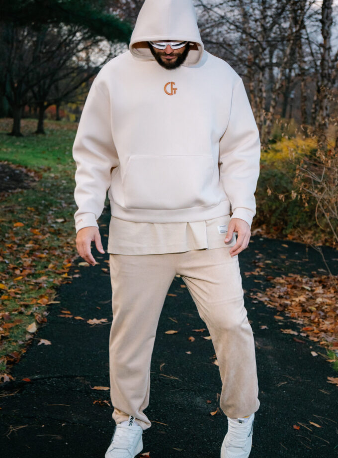 A man wearing a cream-colored hoodie and sweatpants, standing outdoors on a fall evening with trees and fallen leaves in the background, showcasing casual streetwear fashion with a focus on comfort and style.